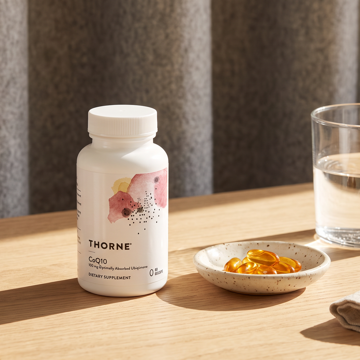 A bottle of THORNE Gato 1000 dietary supplement capsules sits on a light wooden table next to a glass of water and a small dish of orange-yellow capsules, set against a neutral gray curtain background.
